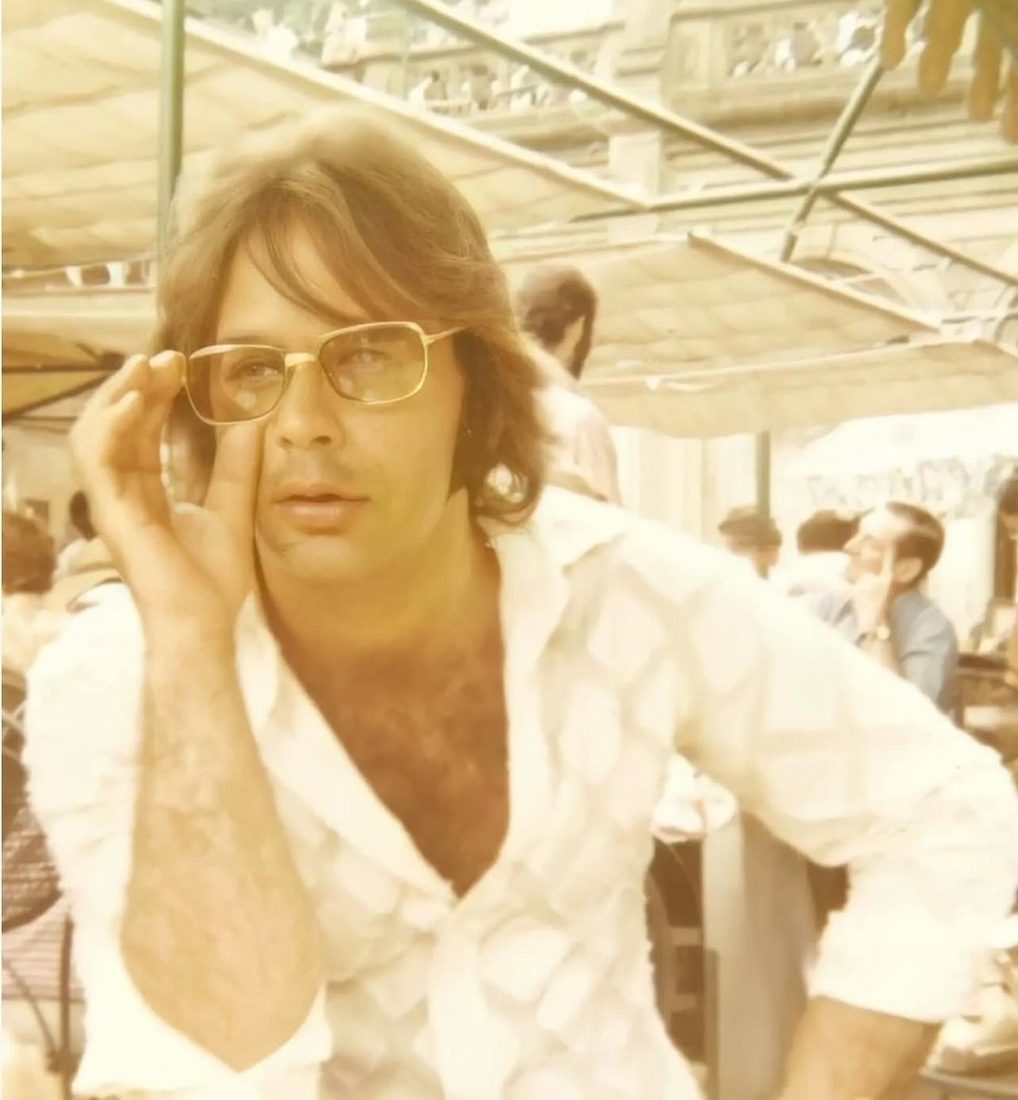 Lou Christie in a white shirt, adjusting his glasses at a café table, 1969